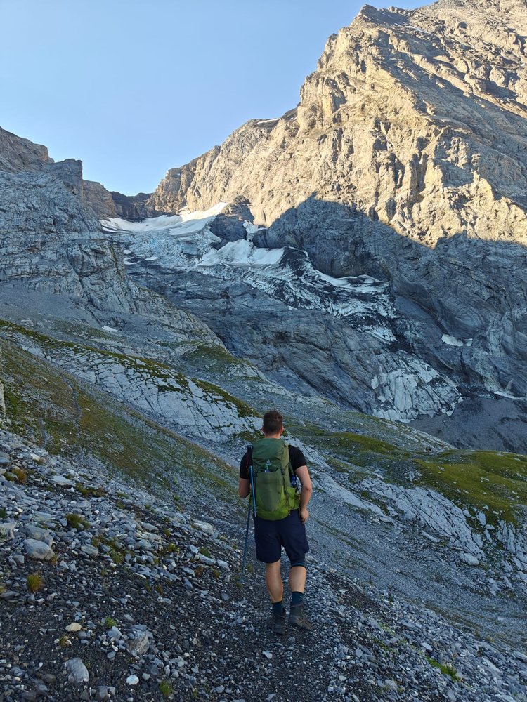 A hiker on a scree path approaching a glacier below steep limestone walls