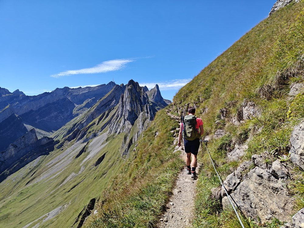 A hiker on a narrow grassy ridge trail with jagged limestone towers ahead